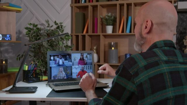 Positive attractive mature man t making video call online on laptop, talking and sharing with multiracial college friends about latest news while enjoying leisure at home at night. Rear view.