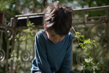 A young boy in a blue shirt is captured outdoors, looking down with a thoughtful expression. The...
