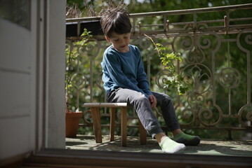 A young boy in a blue shirt sits on a small wooden bench outside on a porch. He looks pensive,...