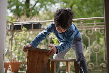 A young boy in a blue shirt, bent over a small wooden bench on a porch, appears to be engrossed in...
