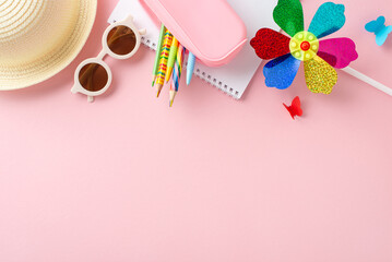 Overhead view of colorful pencils, notebooks, a straw hat, sunglasses, and a decorative pinwheel arranged neatly on a pink background, depicting summer fun and education