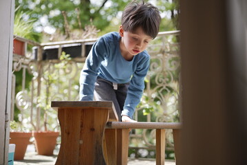 A young boy in a blue shirt leans over a small wooden bench on a porch. His posture and expression...