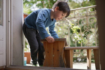 A young boy in a blue shirt climbs onto a small wooden bench on a porch. He appears curious and...