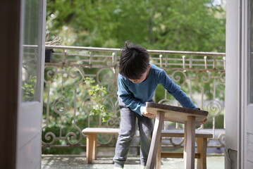 boy sits on a small wooden bench on a balcony, dressed in a blue long-sleeve shirt and grey pants....
