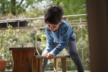 A young boy in a blue shirt is seen bending over a small wooden bench on a porch. He appears intent...