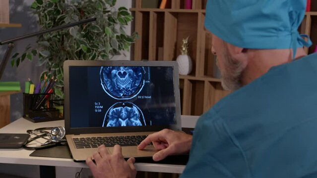 Rear view of middle aged male physician in uniform working on laptop pc, analyzing MRI brain scan of sick patient, thinking about diagnosis and cause of disease at medical office in evening.