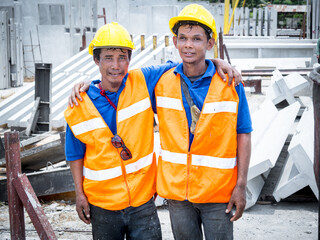 Portrait of two friends Asian workers wearing safety vests with yellow hats hugging together in the precast factory site. People working in construction sites.