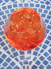 Close-up, high-angle shot of an Aperol Spritz cocktail with ice cubes, glowing in the sunlight, set on a patterned surface