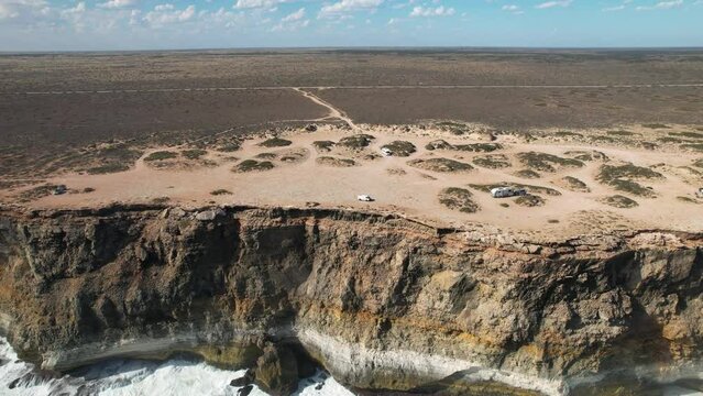 Drone shot flying towards tall cliffs with the waves crashing below. As the drone reaches the cliffs you can see a couple standing near a camper van admiring the view. Bunda Cliffs, South Australia.