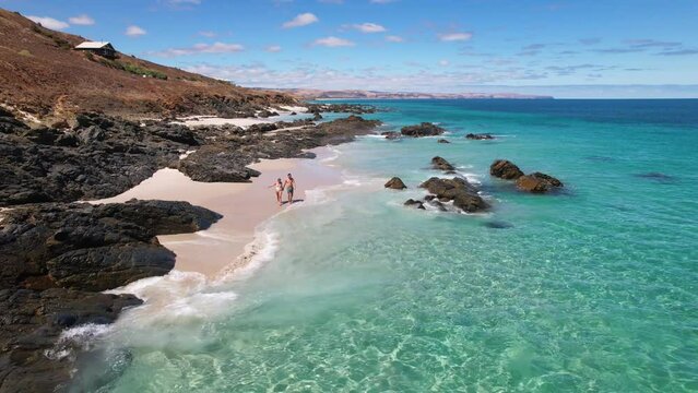 A couple walk hand in hand on a white sand beach into the bright blue crystal clear ocean and start to swim and play while there is no one else in sight. Carrickalinga beach, South Australia.