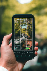 Holding a phone with one hand shooting a vertical video outside in the forest with beautiful river coming down.