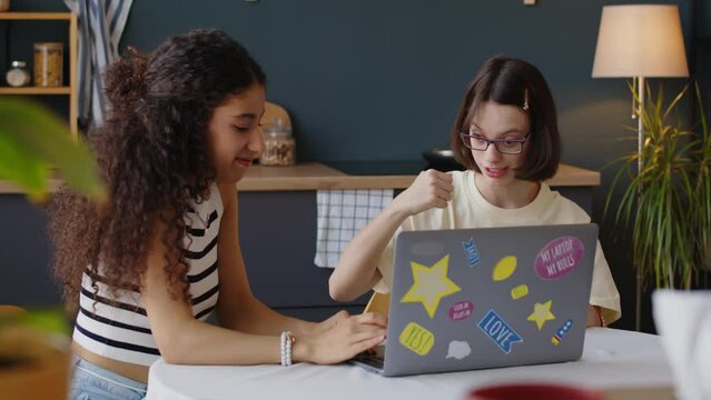 Medium shot of Caucasian teenage girl with cerebral palsy talking to Biracial female friend while studying on laptop together sitting at kitchen table in modern apartment