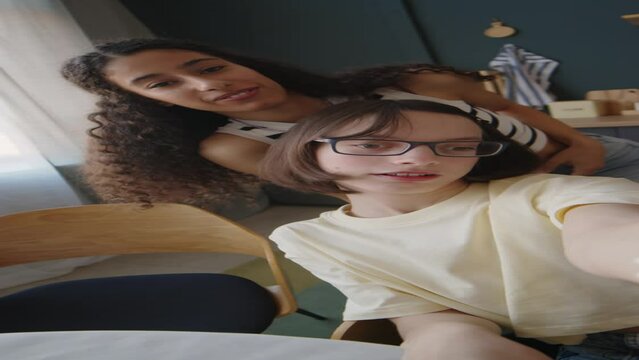 Vertical handheld POV shot of Caucasian teen girl with cerebral party filming video blog with Biracial female best friend spending time together at home