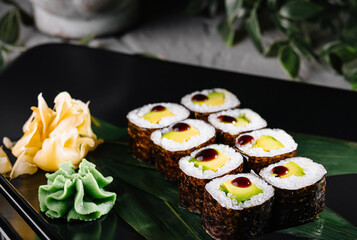Sushi rolls garnished with wasabi and ginger on a modern black plate, botanical backdrop