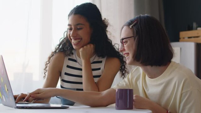 Waist up of Caucasian girl with cerebral palsy with Biracial female best friend using laptop and laughing, sitting at table in modern apartment and spending leisure time together