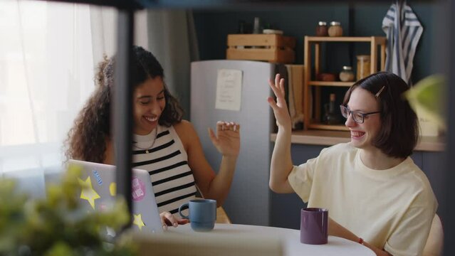 Medium shot of Caucasian teenage girl with cerebral palsy and her Biracial female friend sitting at table and watching funny series on laptop then giving high fives and smiling