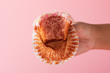 Female hand showing a bitten cupcake in foreground. Pink background.