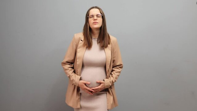 Sick unhappy pregnant woman wearing dress jacket and glasses isolated over gray background feeling start of contractions and strong pain in her belly