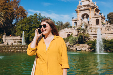 Beautiful stylish woman in trendy wear spending time on street using smartphone. Happy girl talking on the mobile phone near Ciutadella Park in Barcelona. Traveling Europe