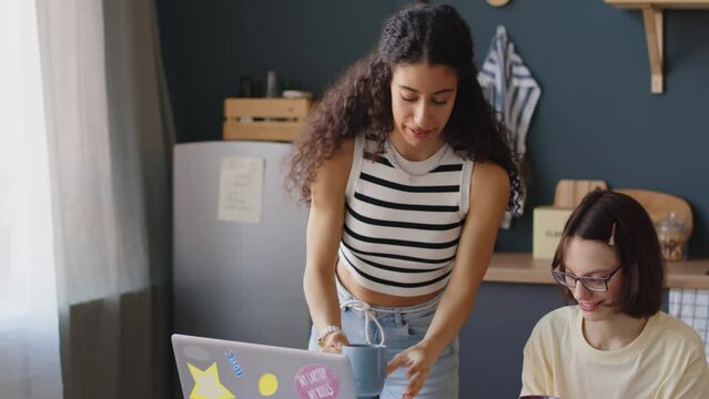 Medium shot of Biracial teenage girl serving two tea cups on kitchen table while spending leisure time with Caucasian female friend with cerebral palsy, watching series on laptop and chatting
