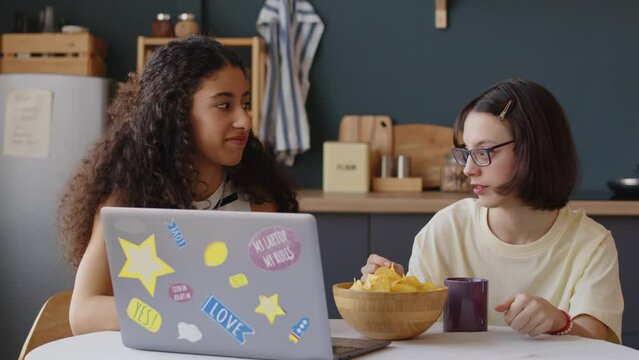 Waist up of Caucasian teenage girl with cerebral palsy eating chips while watching film on laptop with Biracial female best friend, sitting at table in cozy apartment at daytime