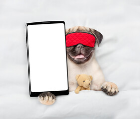 Pug puppy wearing sleeping mask lying on a bed at home and showing big smartphone with white blank...