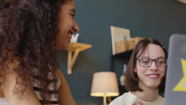 Chest up of Caucasian teenage girl with cerebral palsy talking with Biracial female best friend while watching funny series on laptop at home together