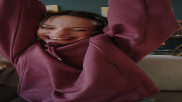 Vertical waist up of Caucasian teen girl with cerebral palsy rejoicing when winning video game, playing on sofa at home