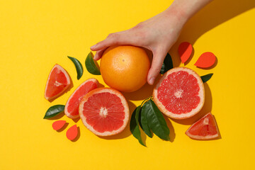 Whole and sliced grapefruit, leaves and female hand on yellow background, top view