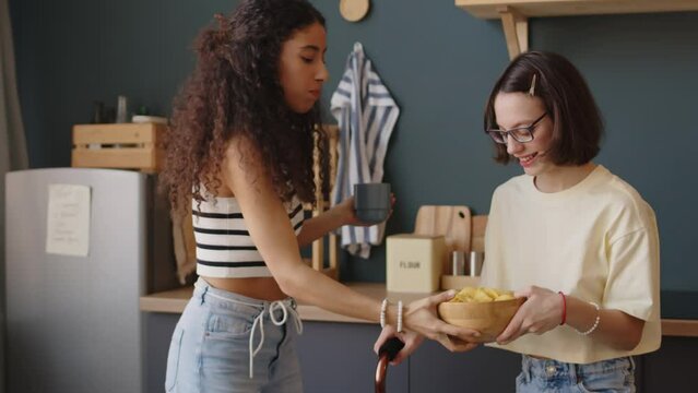 Medium shot of two diverse teenage best female friends eating chips and chatting while spending leisure time together in cozy apartment. One of girls having cerebral palsy
