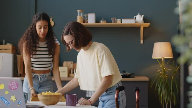 Medium shot of Biracial teenage girl helping Caucasian girl with cerebral palsy to sit down at kitchen table while studying together at home after school
