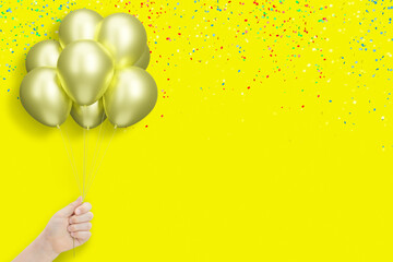 Female hand holding bunch of shiny golden or yellow balloons on yellow background with confetti. Empty space for text
