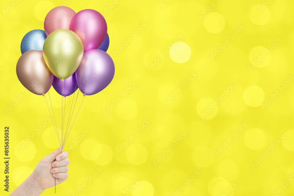 Poster childrens hand holds bunch of colorful shiny balloons on blurred yellow background. empty space for 
