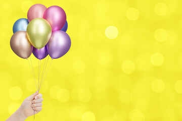 Childrens hand holds bunch of colorful shiny balloons on blurred yellow background. Empty space for text