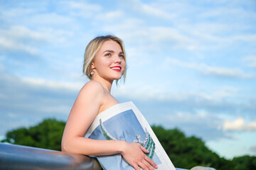 Cute Caucasian Winsome Caucasian Girl Posing Reading Newspaper Against River Outdoors. Horizontal Image
