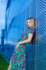 Lifestyle Ideas. Fashion Portrait of Winsome Pretty Cute Girl Woman in Colorful Dress Standing With Lifted Hand in Front of Metal Wall Construction Outdoors.