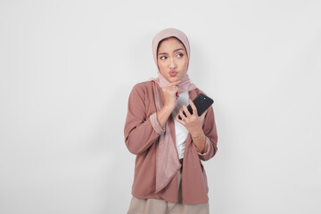 Thoughtful Asian Muslim woman wearing hijab looking aside while holding smartphone isolated over...