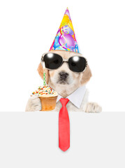 Golden retriever puppy wearing sunglasses, necktie and party cap looks above empty white banner and shows cupcake with burning candle. isolated on white background