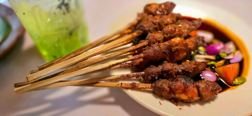 Lamb, mutton grilled skewer satay served with soy sauce sauce with sliced shallots, tomatoes, cucumber and cayenne pepper served in white plate
