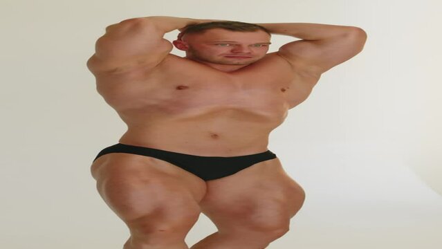 Vertical shot of tanned Caucasian male bodybuilder in perfect shape posing on white background showing muscles while having studio photoshoot indoors