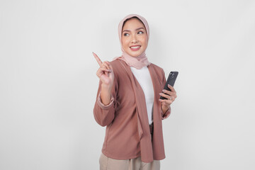 Asian Muslim woman in hijab holding a smart phone while pointing up to the copy space behind over isolated white background.