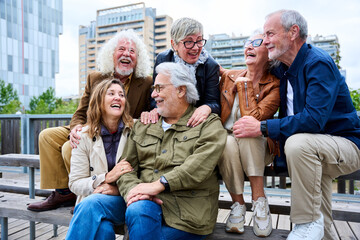 Group of elderly Caucasians laughing together sitting on bench in city. Happy old friends with gray...