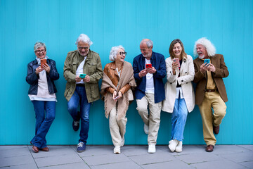 Group of elderly Caucasian laughing together using cell phone standing leaning on blue wall. Happy...
