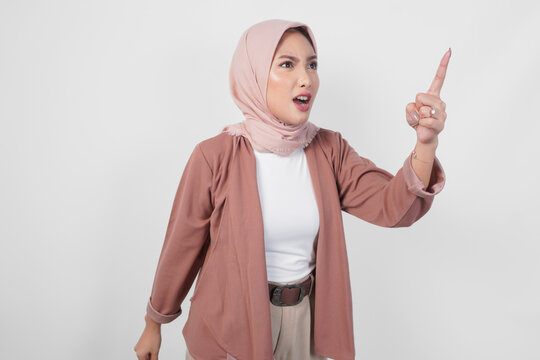 Young Asian Muslim woman wearing hijab looking disgruntled irritated expression while pointing to camera, isolated by white background.