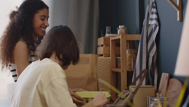 Medium shot of Caucasian girl with cerebral palsy and her Biracial female friend chatting while unpacking paper bags on kitchen table after doing grocery shopping together