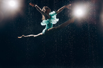 Ballerina teenager girl in green bodysuit bouncing and performing gymnastic splits against falling water drops and spotlight on dark background.