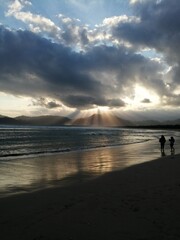 two people are walking on the beach with the sun shining through the clouds