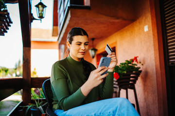 One young caucasian woman is buying or paying online on her mobile phone using credit card on her balcony 
