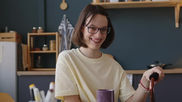 Waist up slowmo portrait of positive Caucasian teenage girl with cerebral palsy sitting at kitchen table in cozy apartment and smiling at camera