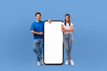 A cheerful young couple standing side by side, confidently presenting a large, blank mobile phone...